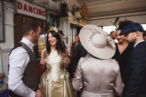 A group of well-dressed people are gathered at an indoor social event. A woman in a gold dress and crown is smiling and holding a drink. Others are engaged in conversation, with one wearing a large hat. A Dancing sign is visible on the wall, capturing the charm that Bristol wedding photographers adore.