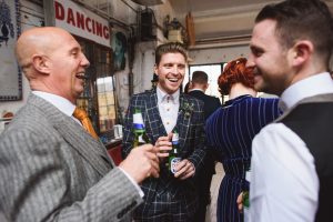 Three men in suits stand together, smiling and holding beer bottles. One wears a checkered suit, another a gray suit with an orange tie, and the third a gray vest. Captured by Somerset wedding photographers, a Dancing sign is visible in the background—a perfect moment of celebration.