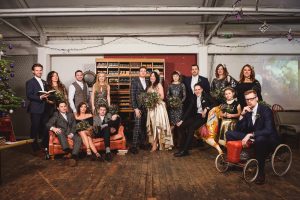 A group of people dressed in formal attire pose in an industrial-style room with wooden floors. Some are sitting on a couch, others stand holding glasses and bouquets. Captured by Bristol wedding photographers, a man is seated on a toy car in the foreground, near a Christmas tree on the left.