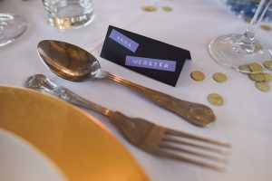 A place setting on a white tablecloth features ornate silverware, a gold-rimmed plate, and a glass. A black name card with Anna Webster in white and purple text sits by the spoon. Gold confetti is scattered around, ready for Bristol wedding photographers to capture this elegant moment.