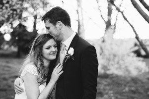 A couple, captured by documentary wedding photographers, embraces outdoors in a black and white photo. The man, wearing a suit with a boutonniere, kisses the womans forehead. She smiles softly while leaning into him, surrounded by blurred trees and grass.