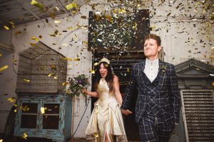 A bride in a cream dress holds a bouquet while the groom in a checkered suit walks hand in hand with her, smiling energetically. Gold confetti falls around them in a vintage room with industrial decor, captured beautifully by Somerset wedding photographers.