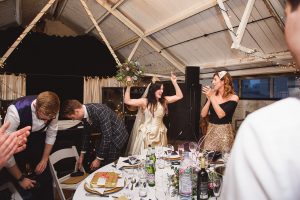 A joyous bride holds a bouquet aloft, celebrating with guests at the reception. One woman claps, while a man adjusts his jacket. The table is set with flowers, glasses, and bottles under a string-lit ceiling in this rustic venue captured beautifully by Bristol wedding photographers.