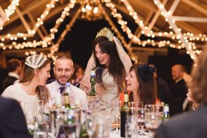 A joyful bride in a white dress and veil with a gold crown leans over to chat with guests at a table. Surrounding her are festively dressed attendees, captured beautifully by skilled wedding photographers from Bristol, under strings of warm lights and a rustic wooden ceiling.