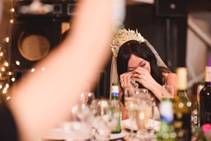 A woman wearing a crown and veil laughs while sitting at a table with drinks and glassware. Captured by documentary wedding photographers, she holds a tissue to her face amidst the celebration, possibly a wedding, with blurred lights twinkling softly in the background.