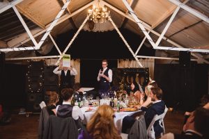 A person stands at a microphone, reading a card, while another holds up a large box with Quirk written on it. The room, reminiscent of the elegant venues favored by wedding photographers in Bristol, is adorned with string lights and a chandelier as guests watch the performance.