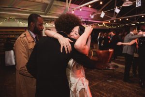 A woman in a white dress hugs a man with curly hair in a formal setting. Captured by Somerset wedding photographers, other elegantly dressed people are visible in the background, with strings of lights adding to the enchanting ambiance.