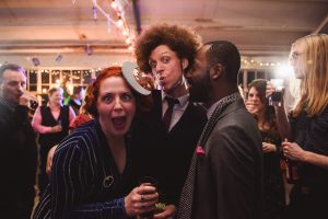 At a lively celebration captured by Somerset wedding photographers, two men and a woman make funny faces. One man with a large afro pretends to kiss through a crescent moon cutout, while the wide-eyed woman and smiling friend revel in the fun. Others enjoy mingling in the vibrant background.