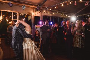 A couple wearing formal attire shares a dance in a warmly lit room decorated with string lights. Documentary wedding photographers capture the moment as guests, including a child, look on, some holding drinks. A DJ booth is partially visible in the background.