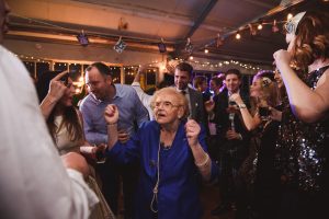 Amidst the lively party, an elderly woman in a blue outfit dances energetically, surrounded by smiling guests and string lights in a festive setting. Its a moment beautifully captured by Somerset wedding photographers, highlighting the joy that fills every corner of the celebration.