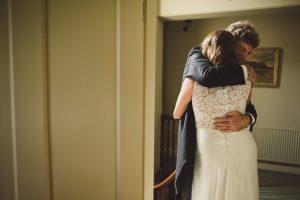 In a warmly lit room, a man and woman share an emotional embrace. She wears a white dress with lace details; hes in a dark suit. With a painting and staircase as their backdrop, the moment is beautifully captured by Somerset wedding photographers.