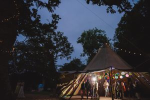 At dusk, a large tent in the wooded area comes alive with vibrant paper lanterns. Inside and outside, people bask in the glow of soft lights. As trees frame this enchanting scene, Bristol wedding photographers capture every magical moment amidst string-lit branches.