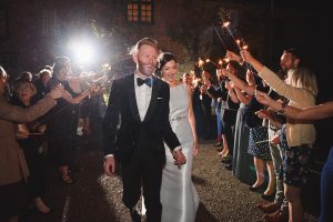 A newlywed couple walks hand in hand through a joyful crowd. Guests hold sparklers, illuminating the night. Captured perfectly by wedding photographers Somerset, the bride in her white gown and the groom in his tuxedo smile blissfully amid the enchanting scene.