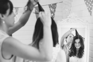 A woman smiles at her reflection in a mirror as another person styles her hair, captured beautifully by Somerset wedding photographers. The room has festive bunting decorations, and the atmosphere is cheerful. The photo is in black and white.