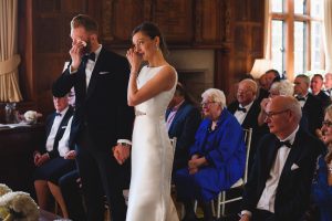A bride and groom stand at the front of a wood-paneled room, both wiping tears. The bride wears a white gown, and the groom is in a black suit. A documentary wedding photographer captures this heartfelt moment as guests, dressed in formal attire, watch the couple with emotion-filled eyes.