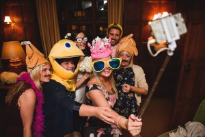 A group of six people in a room are wearing playful hats and oversized sunglasses, reminiscent of a whimsical photoshoot by Somerset wedding photographers. One person is holding a selfie stick, capturing the joyous moment. Their smiles suggest they’re having fun at a lively party or gathering.