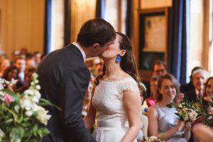 A bride and groom share a kiss at their wedding ceremony, beautifully captured by top Somerset wedding photographers. The bride wears a white gown with lace details, and the groom sports a dark suit. Guests watch from their seats in the warmly lit room, some holding flowers.