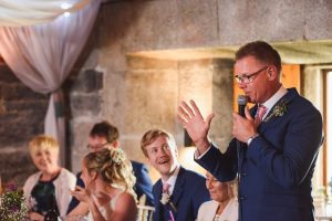 A man in a blue suit is giving a speech with a microphone at the wedding, gesturing passionately. Guests, including a smiling couple, are seated at tables, listening attentively. The warmly lit setting with draped fabric decoration creates the perfect moment for Somerset wedding photographers to capture.