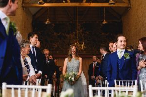 A woman in a sparkling dress walks down the aisle holding a bouquet, smiling. Guests in formal attire watch her. Somerset wedding photographers capture the moment amid wooden beams and a decorated wall, enhancing the venues enchanting allure.