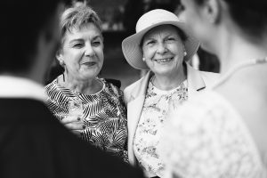 Two older women, stylishly dressed in patterned and floral outfits, are all smiles at a social gathering captured by talented wedding photographers. One holds a drink, while the other sports a wide-brimmed hat. They face someone blurred in the foreground, evoking nostalgia in classic black and white.