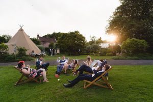 A group of people relax on deck chairs on a lush green lawn during a sunny day, captured beautifully by Somerset wedding photographers. A large tipi tent and trees are in the background, with the sun setting on the horizon. Everyone appears to be enjoying the peaceful outdoor setting.