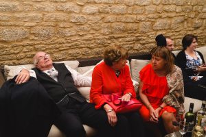 A man in a black vest and white shirt is sleeping on a couch, perhaps exhausted from coordinating with Somerset wedding photographers. Two women in red outfits sit next to him, engaged in conversation. The scene has a rustic stone wall backdrop, with several people gathered in the background.