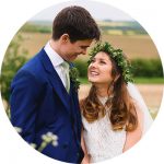 A bride and groom smiling at each other outdoors, captured perfectly by skilled wedding photographers in Somerset. The bride wears a floral crown and a white lace dress, while the groom is in a blue suit with a white tie against the scenic countryside backdrop.