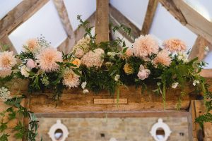 A rustic wooden beam adorned with lush greenery and soft pink flowers, including roses and dahlias. A small sign on the beam reads MIND YOUR HEAD. The room, often captured by Somerset wedding photographers, features wooden beams and stone walls with circular windows in the background.