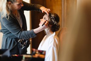 A makeup artist applies eyeliner to a seated bride in a white robe, captured beautifully by Bristol wedding photographers. The bride adorns a detailed floral hairpiece. This intimate indoor scene with soft lighting focuses on preparation for her special day.