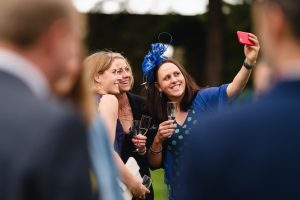 Three women in elegant dresses and a fascinator take a selfie outdoors, champagne glasses in hand and smiles broad. Blurred figures hint at a joyful social gathering, possibly captured by Bristol wedding photographers documenting the joyous occasion.