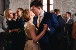 A couple in formal attire kisses and dances at an indoor event, captured perfectly by a talented wedding photographer from the Cotswolds. The woman wears a light-colored dress, and the man is in a dark suit with a patterned tie. Other guests mingle near large windows in the background.