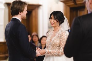 A bride and groom exchange vows indoors. The bride, in a lace wedding dress and veil, gazes at the groom in his dark suit. Guests are seated in the background as a Bristol wedding photographer captures the moment.