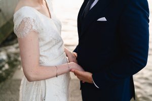 A couple stands closely together near the water, holding hands
