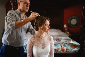 A bride in a lace wedding dress sits on a bed, her hair being styled meticulously by a Somerset wedding photographer. The bedroom, with its warm-toned walls and colorful patterned bedspread, sets a charming scene as the stylist focuses intently on perfecting the bride’s hairstyle.