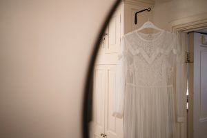 A delicate white lace wedding dress hangs on a wooden hanger in a softly lit room, perfectly poised for a wedding photographer from the Cotswolds to capture its intricate lace details and sheer long sleeves. Its elegance is reflected in a circular mirror on the left.