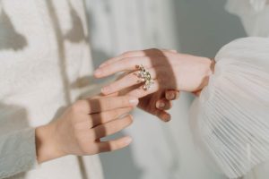 Close-up of two hands; one adorned with a detailed floral ring, gently captured by expert wedding photographers in Bristol. The other hand appears to softly touch it, both wearing light, textured clothing. Soft shadows are cast over them, creating a serene atmosphere.