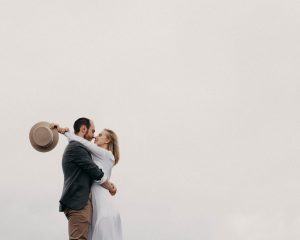 A couple embraces lovingly against a pale, overcast sky. The man holds a brown hat, while the woman, wearing a flowing white dress, wraps her arms around him. Captured by talented wedding photographers in Somerset, this moment reflects timeless romance.