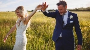 A couple joyfully walks through a sunlit field, hand in hand. The woman wears a white lace dress, while the man is in a blue suit with a white boutonniere. As Somerset wedding photographers capture the moment, yellow flowers dot the grassy field beneath a clear sky.